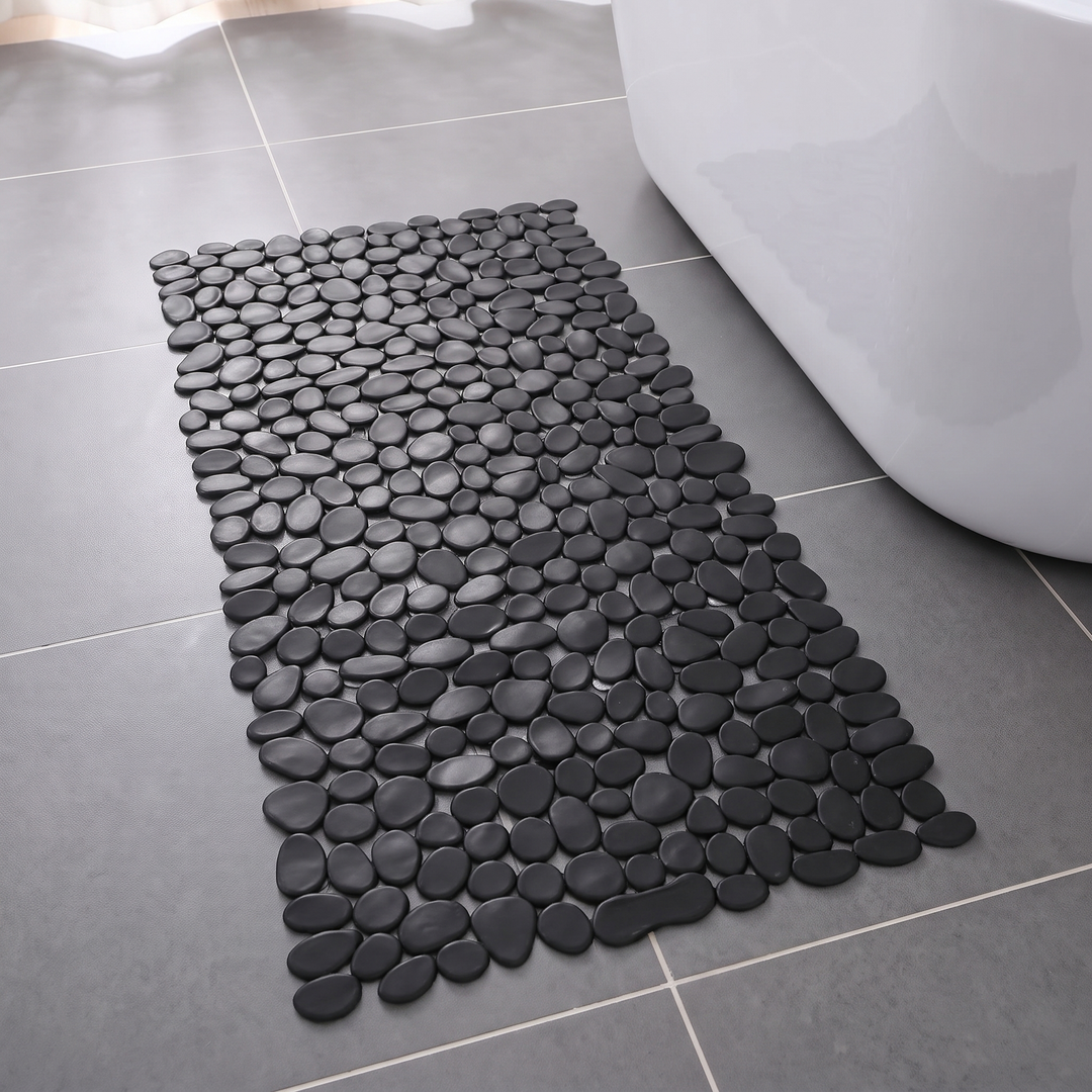 Black pebble shower mat on a grey tiled bathroom floor beside a bath