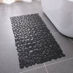 Black pebble shower mat on a grey tiled bathroom floor beside a bath