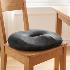 Dark grey donut cushion with its open centre sitting on the seat of an oak dining chair in a bright room by a window