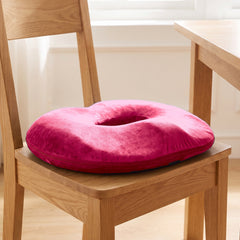 Rose red donut cushion with its open centre sitting on the seat of an oak dining chair in a bright room by a window