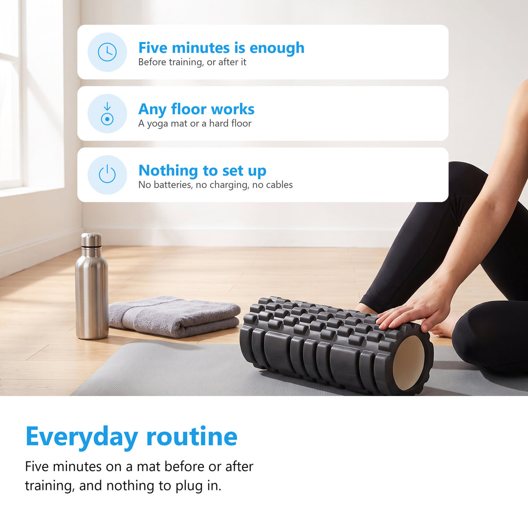 Black textured foam roller on a grey yoga mat in a bright living room with a water bottle and a towel