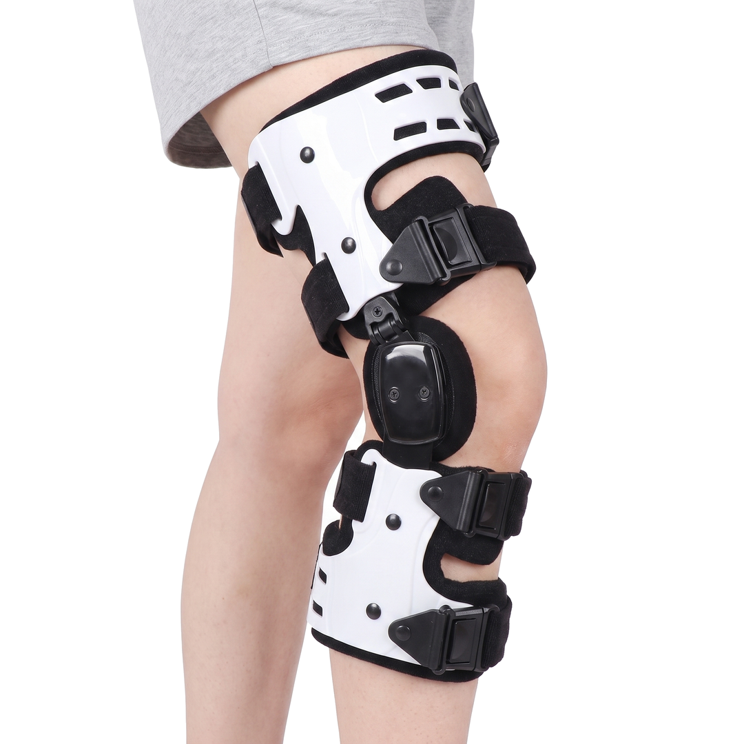 A woman standing on a pure white background wearing a white hinged knee brace with two rigid shells, a side hinge and four black quick release straps