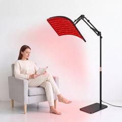 Woman sitting in an armchair beside a red light therapy lamp with a dual wavelength LED panel on an adjustable black floor stand