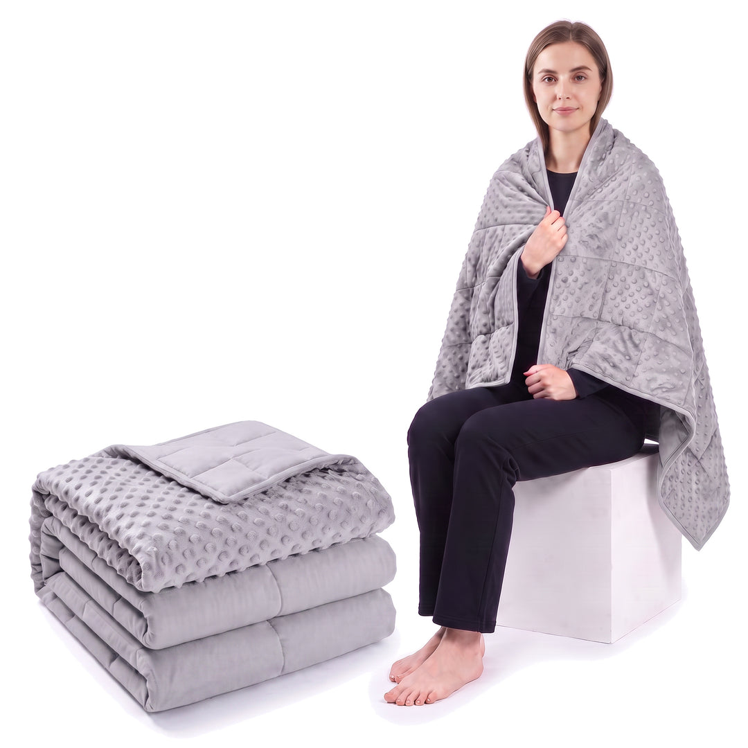 Light grey weighted blanket folded on a white background beside a woman wrapped in the same blanket
