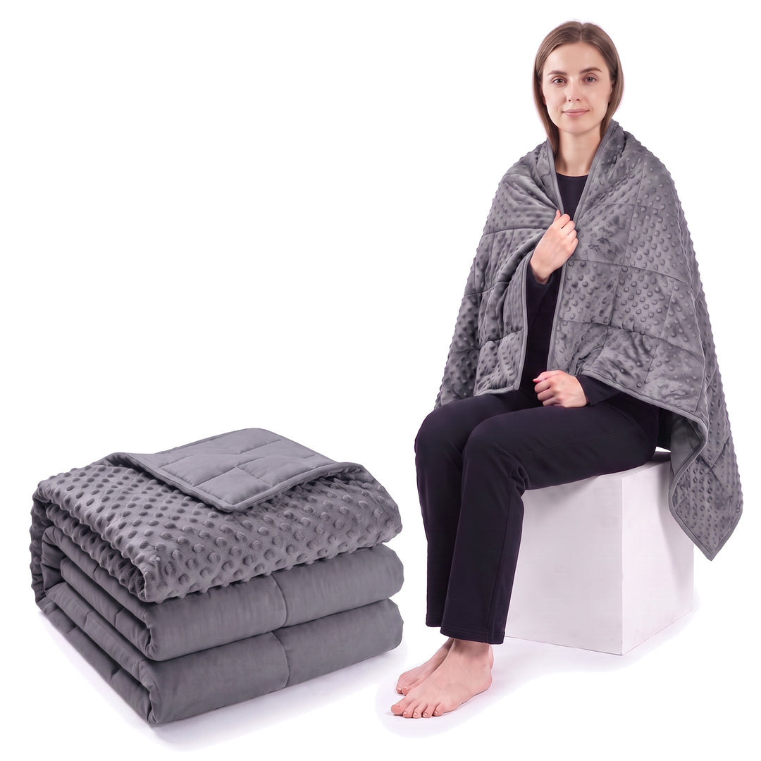 Dark grey weighted blanket folded on a white background beside a woman wrapped in the same blanket